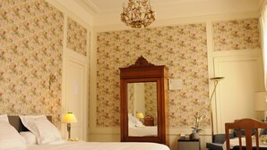 A hotel room with floral wallpaper, a large mirror, a bed with white linens, a nightstand with a lamp, and a chair. - Fine Fleur Bed & Breakfast (Bruges)
