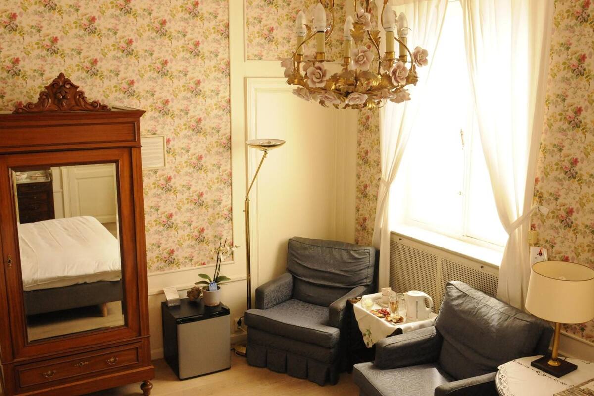 A bedroom with a wooden wardrobe, a chair, a small table with a lamp, and a window with curtains.