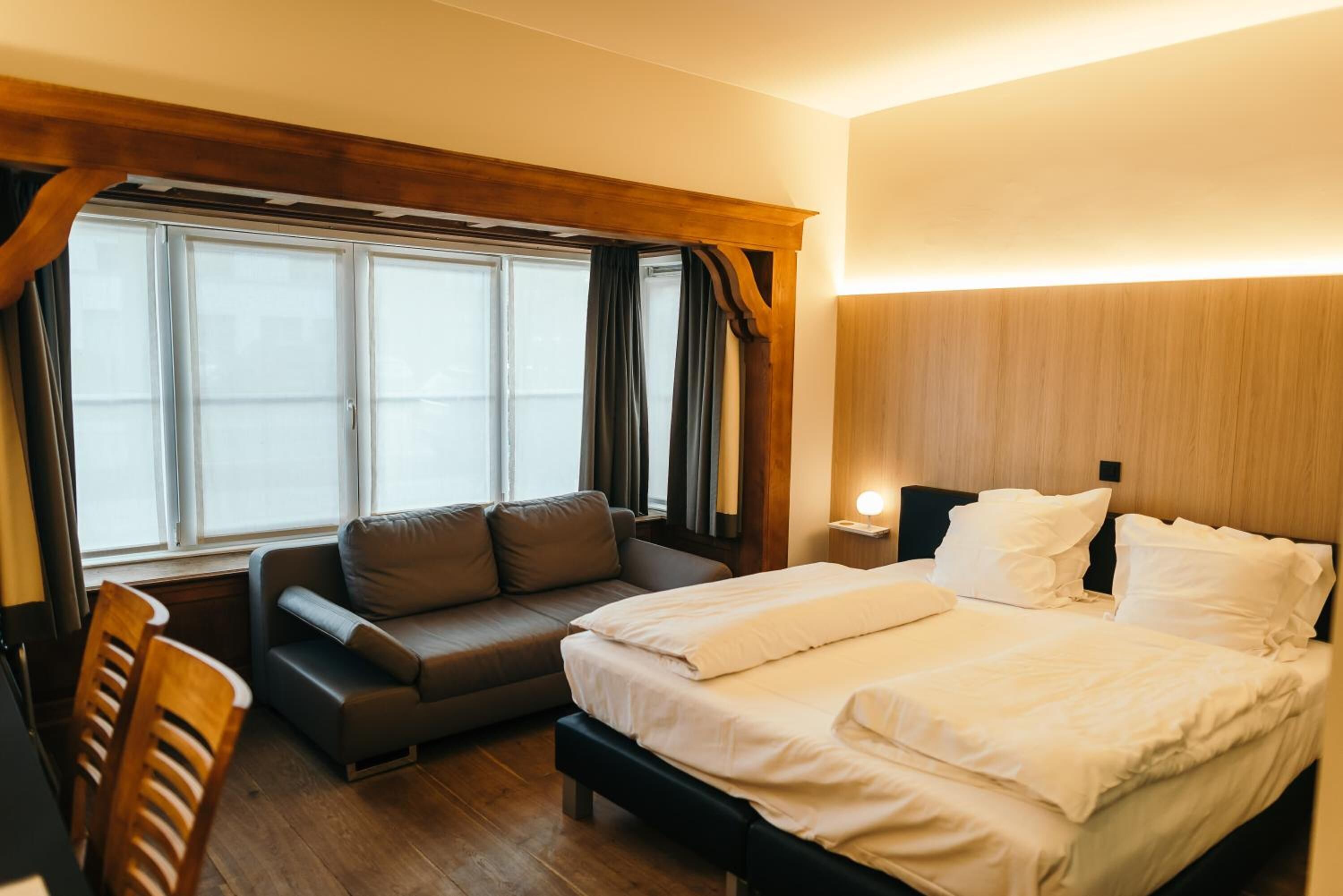 A bedroom with a bed, a sofa, a wooden bench, a nightstand, and a window with curtains.