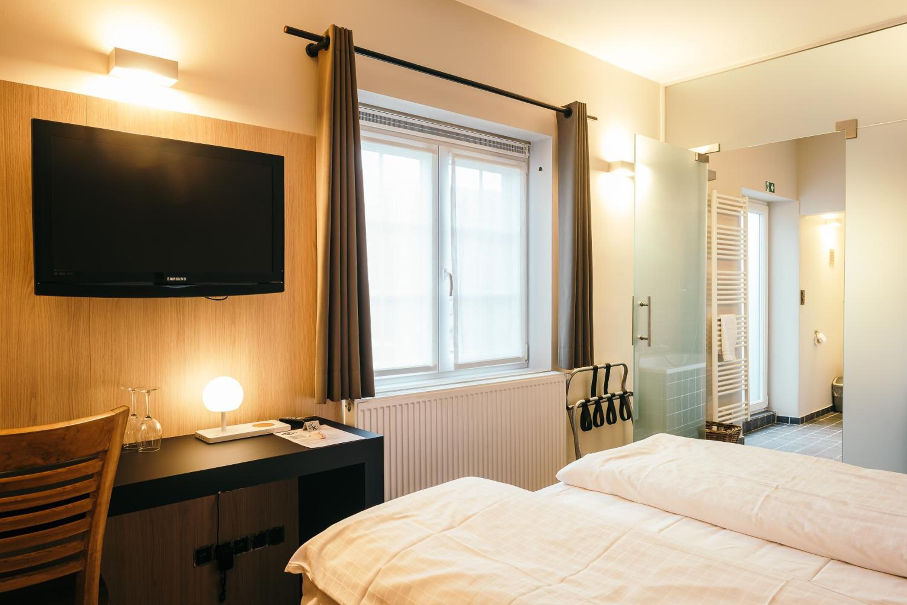 A hotel room with a flat-screen TV mounted on the wall, a desk with a lamp, a bed with white bedding, a window with curtains, and a shower area with a glass door.