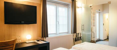Une chambre d’hôtel dotée d’un téléviseur à écran plat fixé au mur, d’un bureau avec une lampe, d’un lit avec une literie blanche, d’une fenêtre avec des rideaux et d’un espace douche avec une porte en verre.