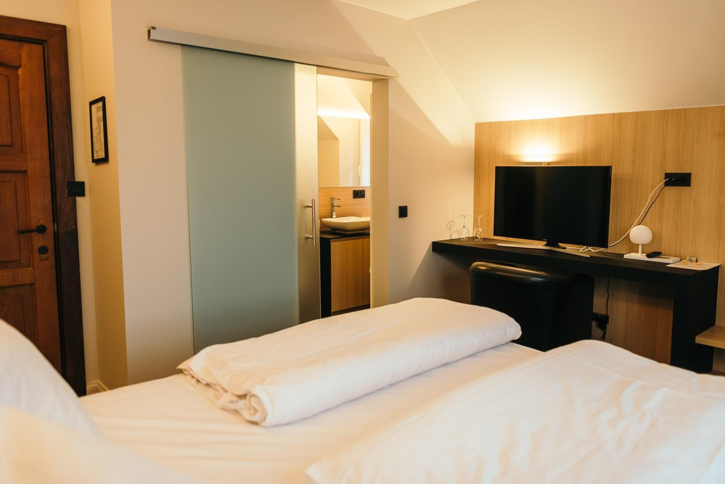 A hotel room with two beds, a TV, a desk, and a bathroom visible through a sliding door.
