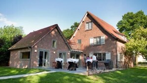 Une maison à deux étages, construite en briques, avec un toit en pente, une terrasse couverte et un petit balcon.