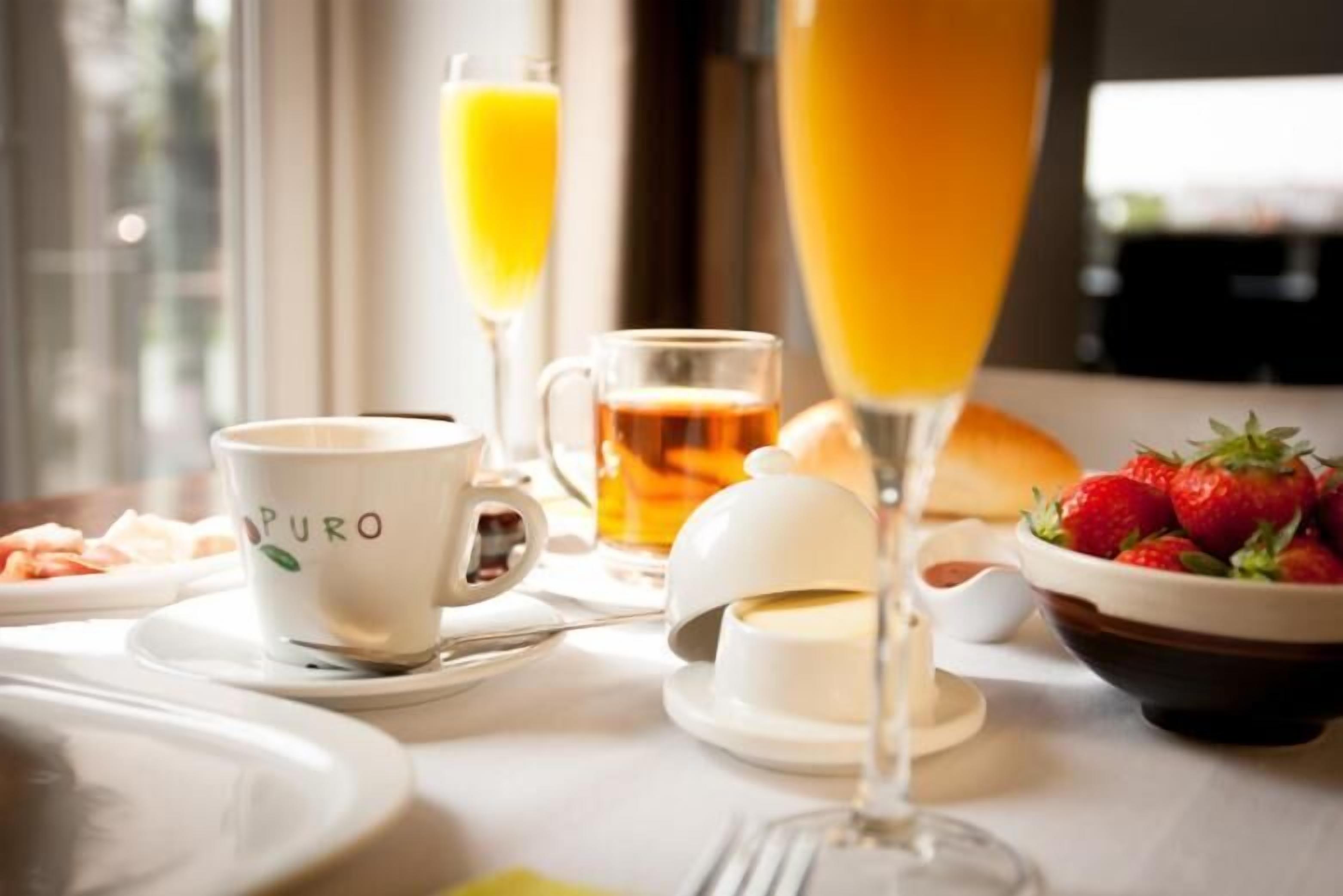 Ein Frühstücksarrangement mit einer Tasse mit dem Aufdruck "PURO", einem Glas Orangensaft, einem Glas Champagner, einer Schale Erdbeeren und einem Teller Croissants.