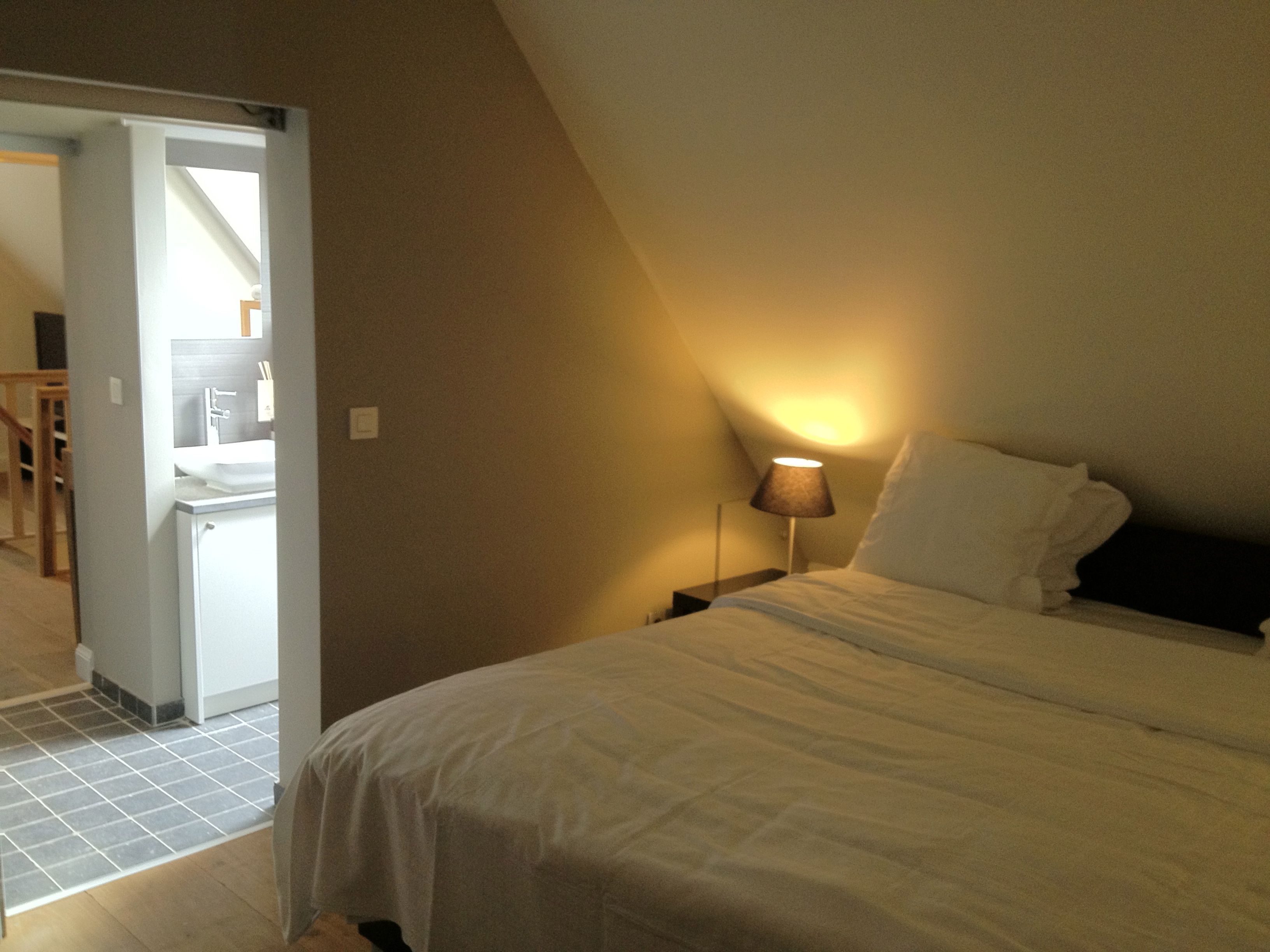 A bedroom with a bed, a nightstand with a lamp, and a bathroom visible through an open door.