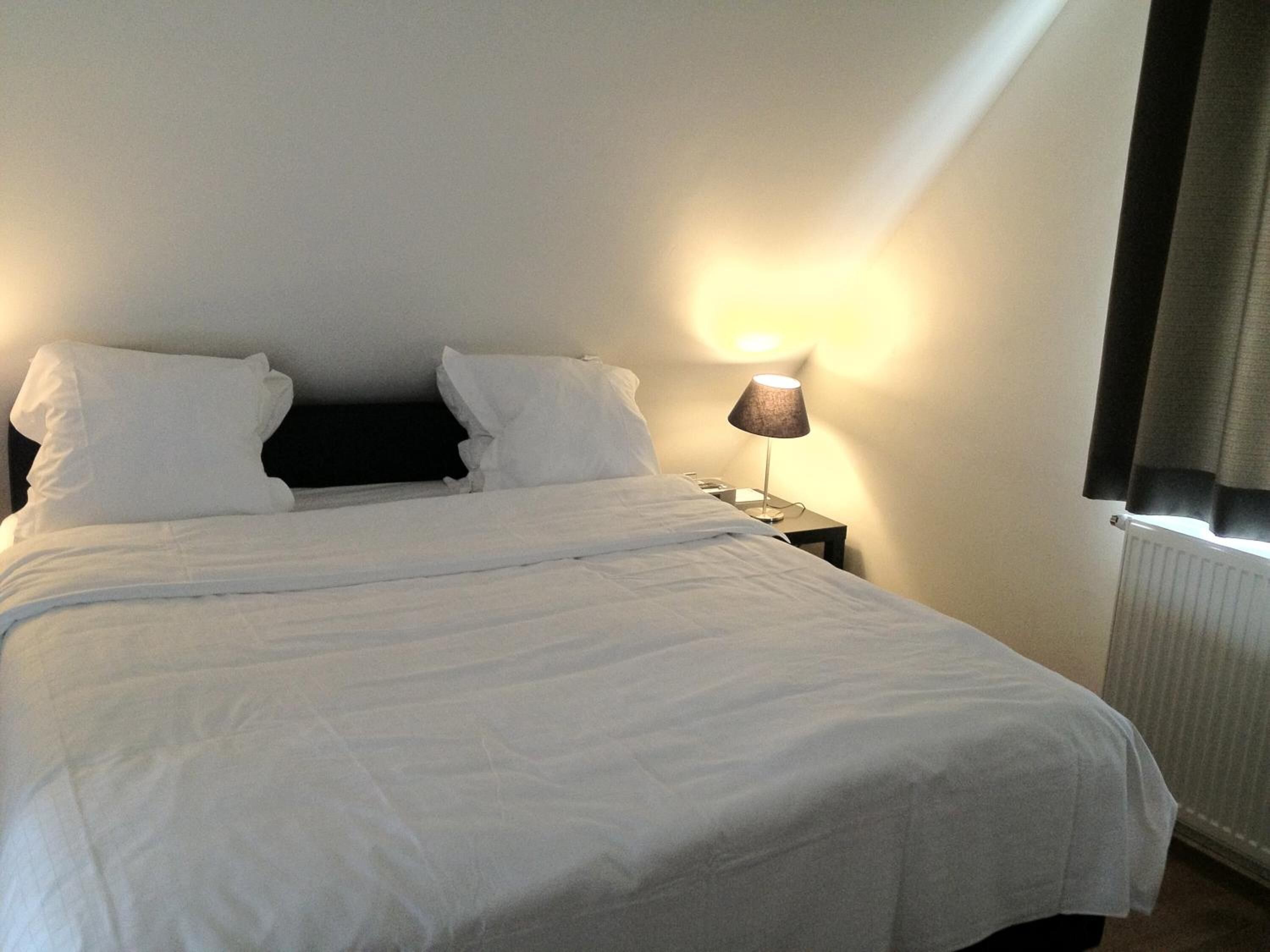 Un lit bien fait, avec du linge de lit blanc, une lampe de chevet et une fenêtre avec des rideaux.