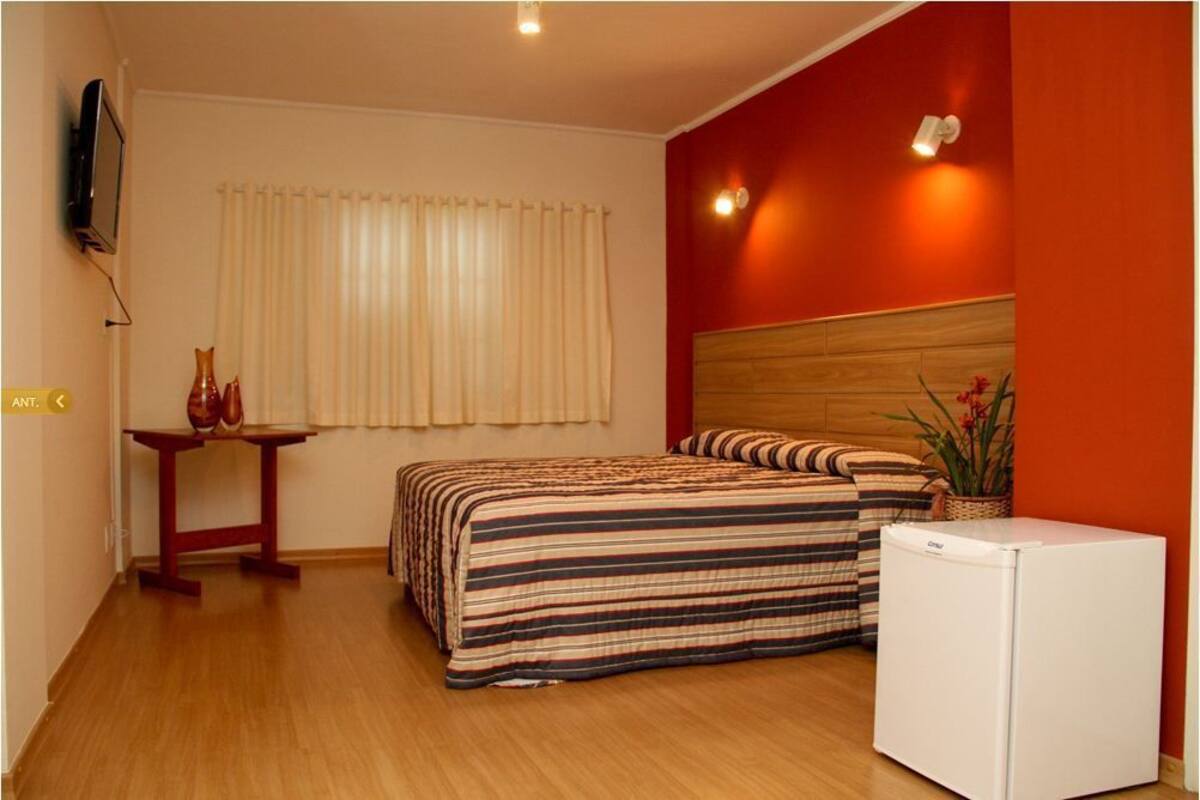 Ein Schlafzimmer mit einem Bett, einem kleinen Tisch mit zwei Vasen, einem Fernseher an der Wand und einem Kühlschrank.
