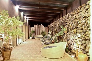 Uma área de estar ao ar livre coberta, com cadeiras de vime e um banco trançado, rodeada por plantas em vasos e um muro de pedra.