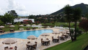 A pool area with lounge chairs and umbrellas, surrounded by a landscaped garden and a view of a lake and hills. - Hotel Nascentes da Serra (Poços de Caldas)