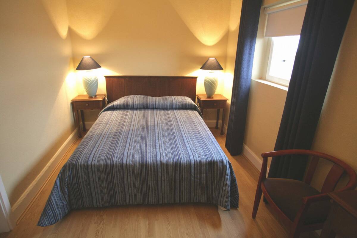 A bedroom with a bed, two bedside lamps, a wooden nightstand, a chair, and a window with curtains.