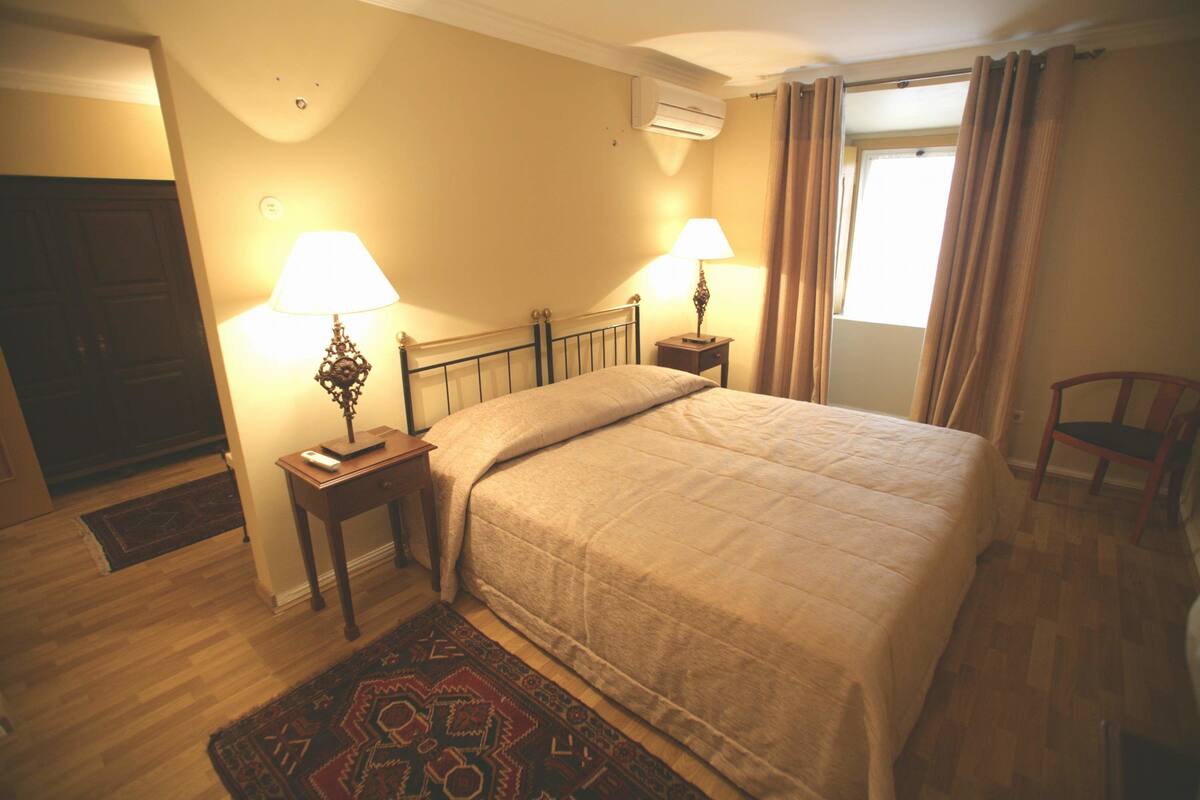 A bedroom with a bed, two bedside tables with lamps, a window with curtains, and a wooden floor.