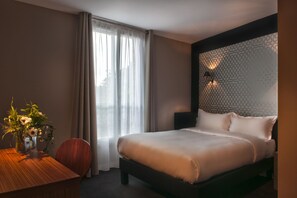 A hotel room with a bed, a desk with a vase of flowers, a chair, and a window with curtains. - Hotel Les Terres Blanches (Chatou)