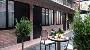 Un área de estar al aire libre con una mesa y sillas, un pequeño jardín con plantas, y un edificio de ladrillo con ventanas grandes y un balcón.