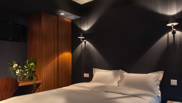 Un dormitorio moderno con cama, paneles de madera, una lámpara de pared y una pequeña mesa con flores.