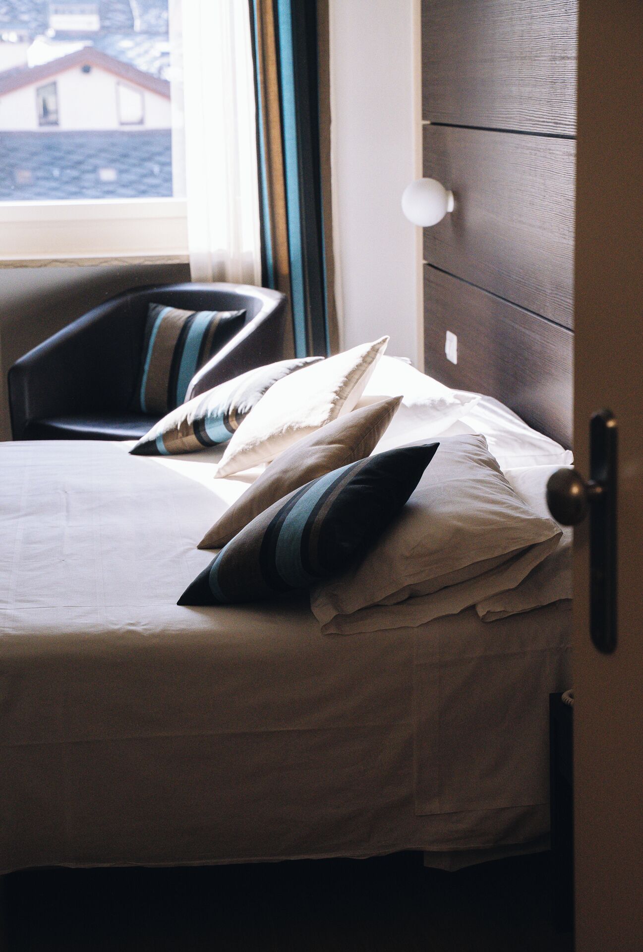 Una cama con una colcha blanca y varias almohadas, un armario de madera y una ventana con vistas a edificios.