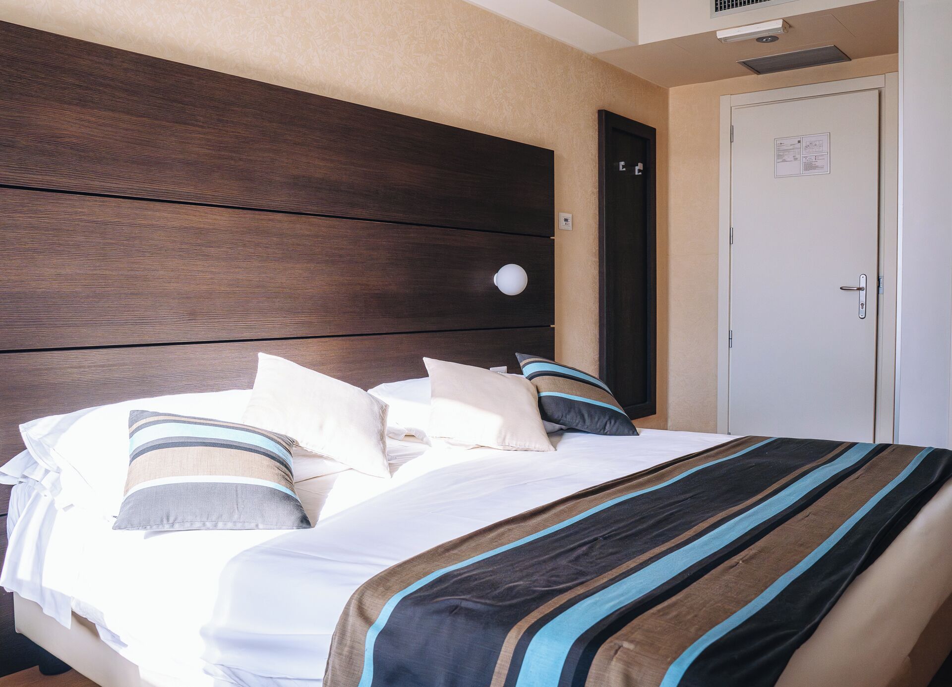 Una habitación de hotel con cabecera de madera, cama con ropa de cama a rayas, una puerta blanca y un interruptor de luz en la pared.
