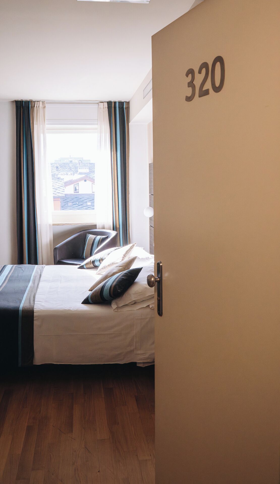 A hotel room with a bed, curtains, a view of buildings outside, and a door with the number 320.