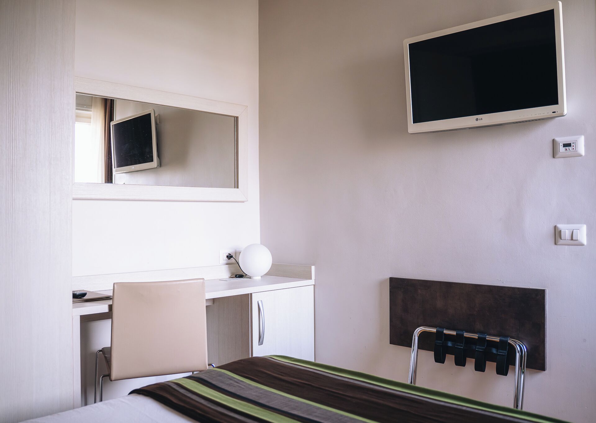 A hotel room with a bed, a desk with a chair, a TV mounted on the wall, and a small bedside table with a lamp.