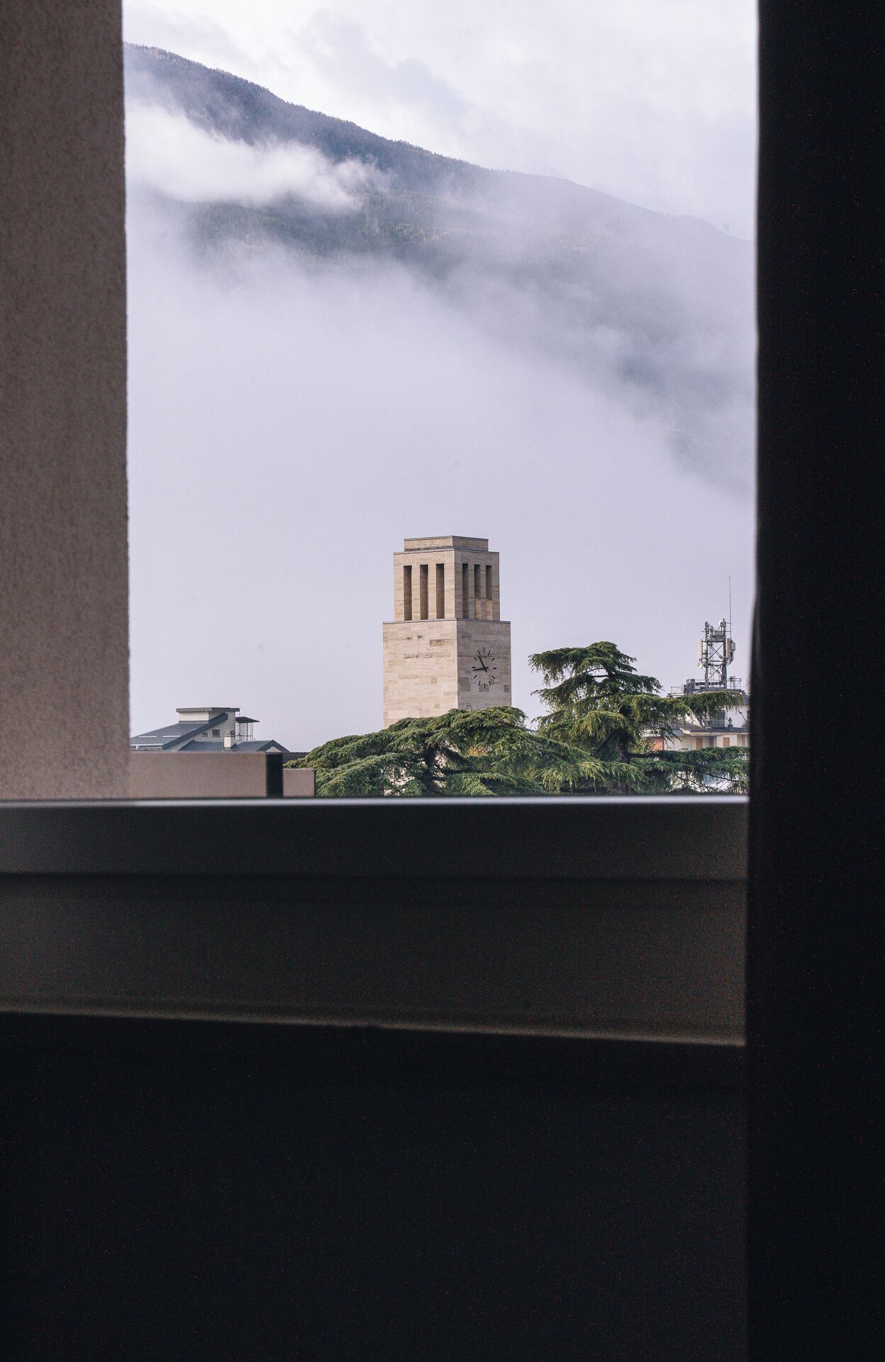 Vista de una torre del reloj con un edificio moderno en primer plano, rodeado de árboles y montañas cubiertas de niebla.
