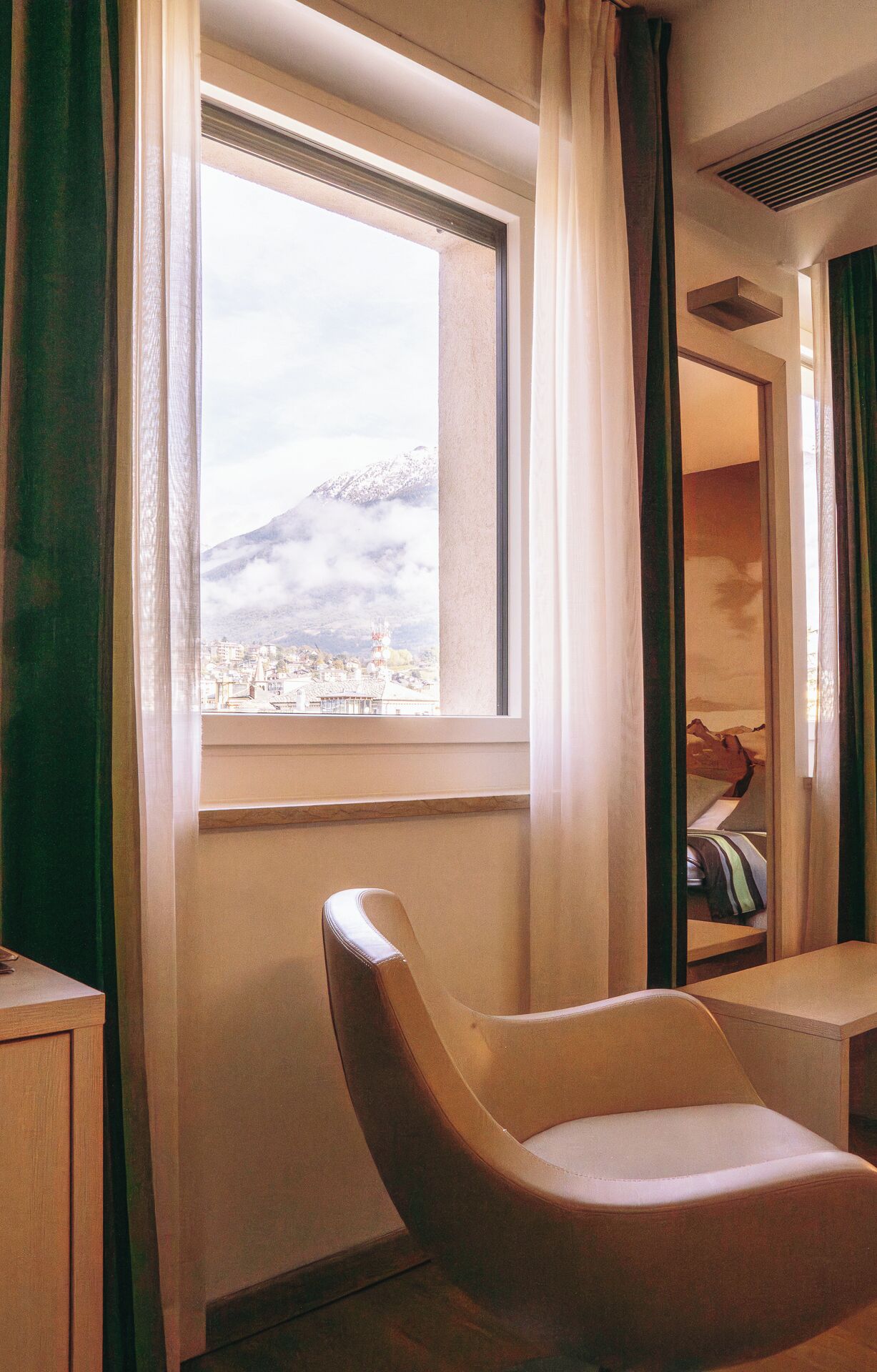 A room with a view of a mountain and cityscape through a window, a chair, and a bed with a striped blanket.