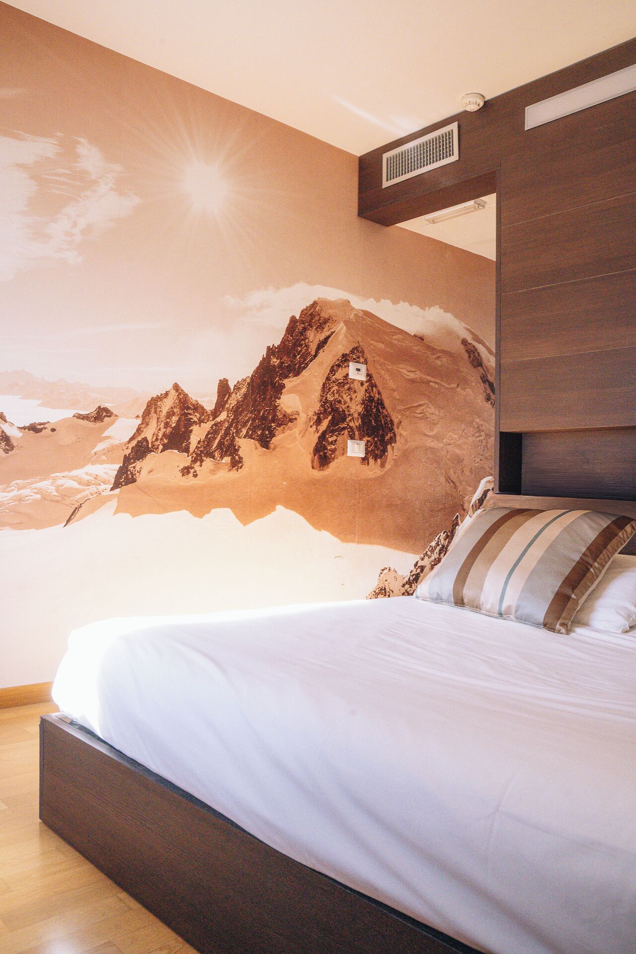 A bedroom with a mountain mural on the wall, a bed with white bedding, and a wooden headboard.