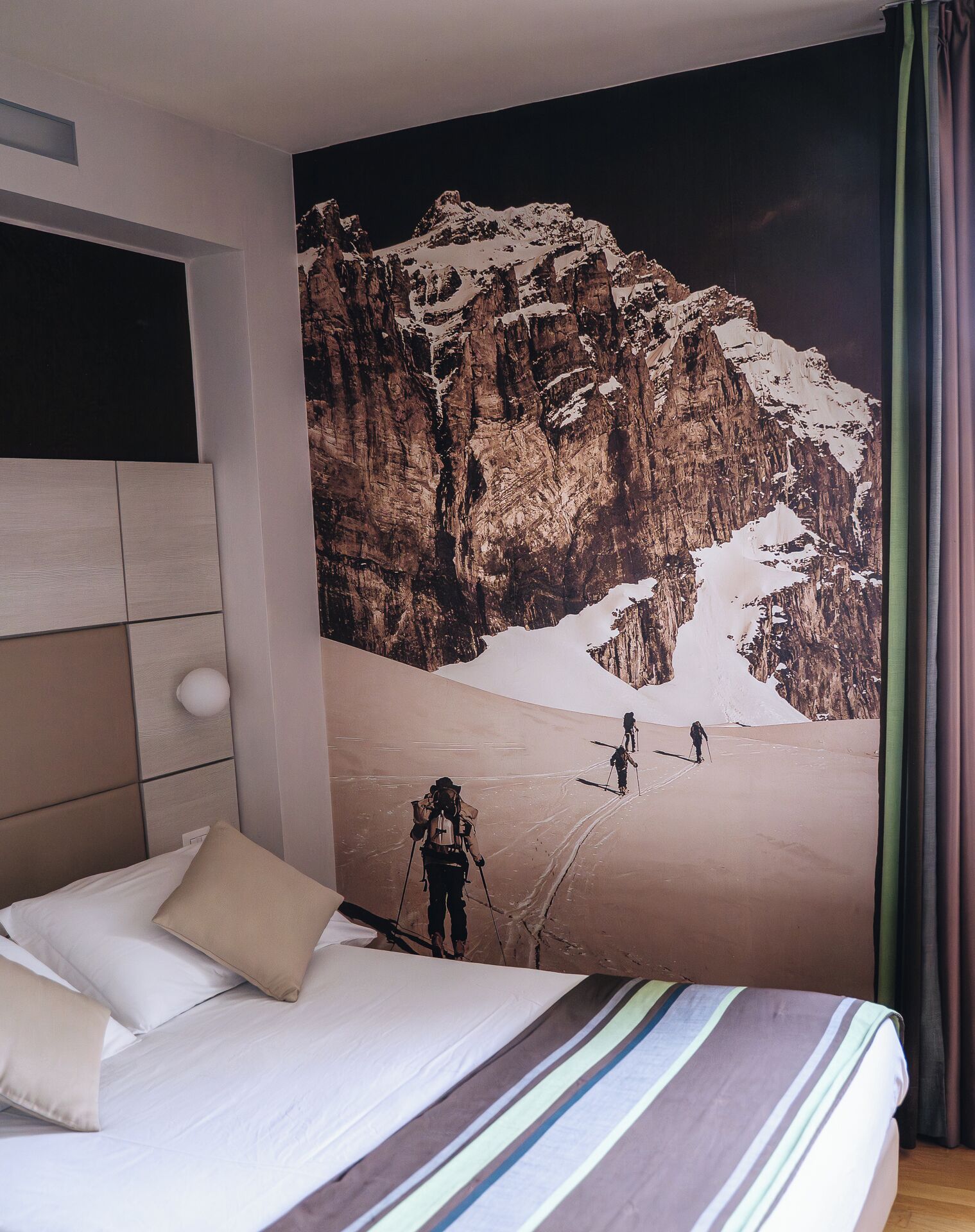 Un dormitorio con un gran mural de un paisaje montañoso en la pared, una cama con ropa de cama a rayas y un cabecero con luz integrada.