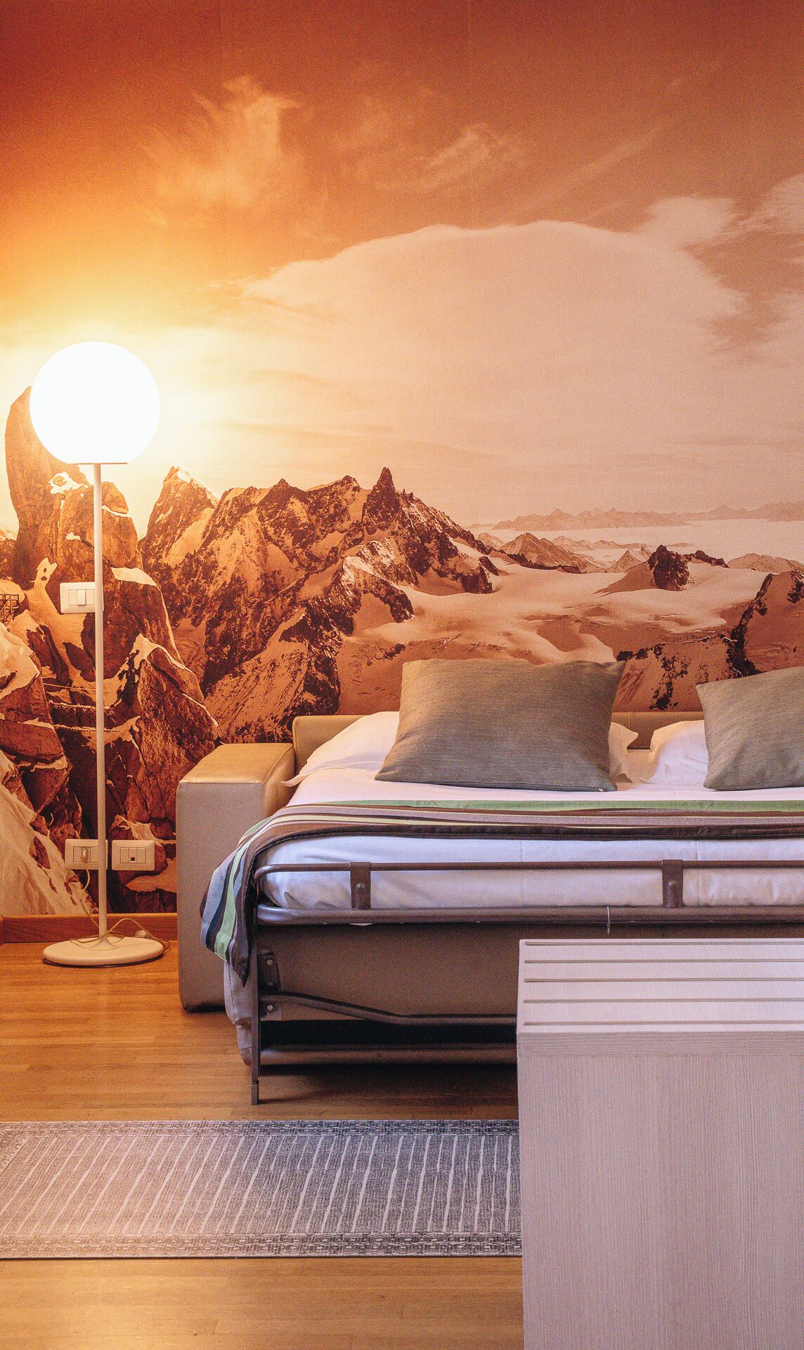 Un dormitorio con un mural de paisaje montañoso, una lámpara de noche y una cama con almohadas.
