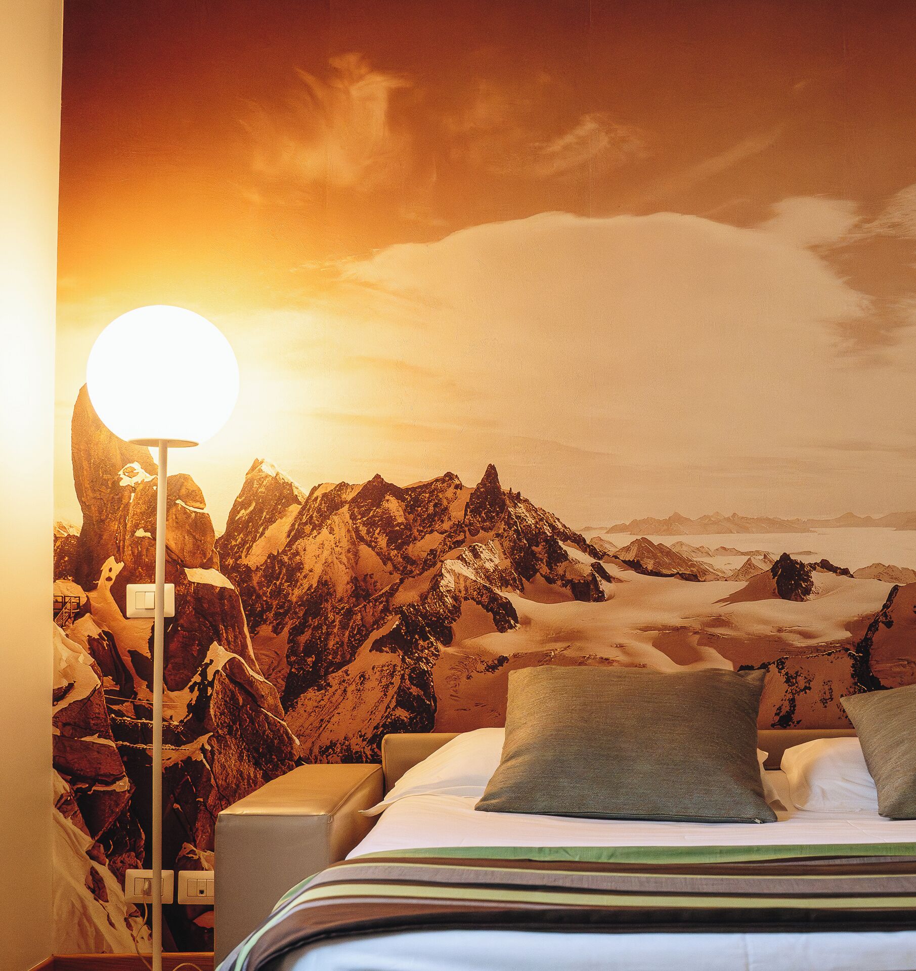 A bedroom with a mountain landscape mural, a bed with pillows, and a bedside lamp.