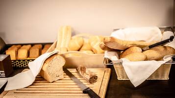 A wooden cutting board with a knife, a loaf of bread, and a basket of baguettes on a dark wooden table.