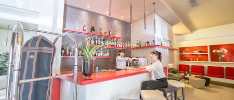 A modern bar area with a red counter, shelves with bottles, and a seating area with red cushions.