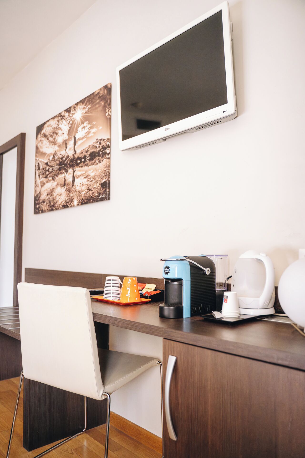 Una camera d'albergo con una scrivania in legno, una sedia bianca, una macchina per il caffè, un bollitore e un quadro appeso al muro.