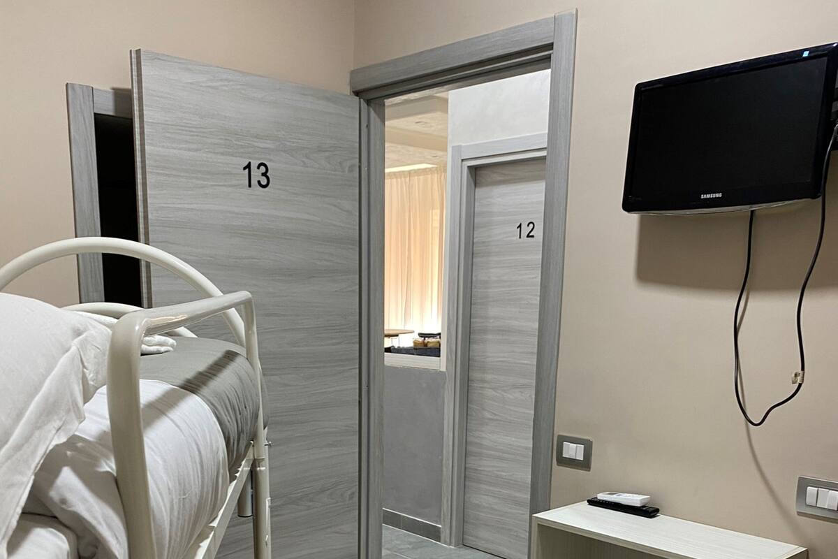 A hotel room with a bed, a TV mounted on the wall, a small shelf, and a trash bin.