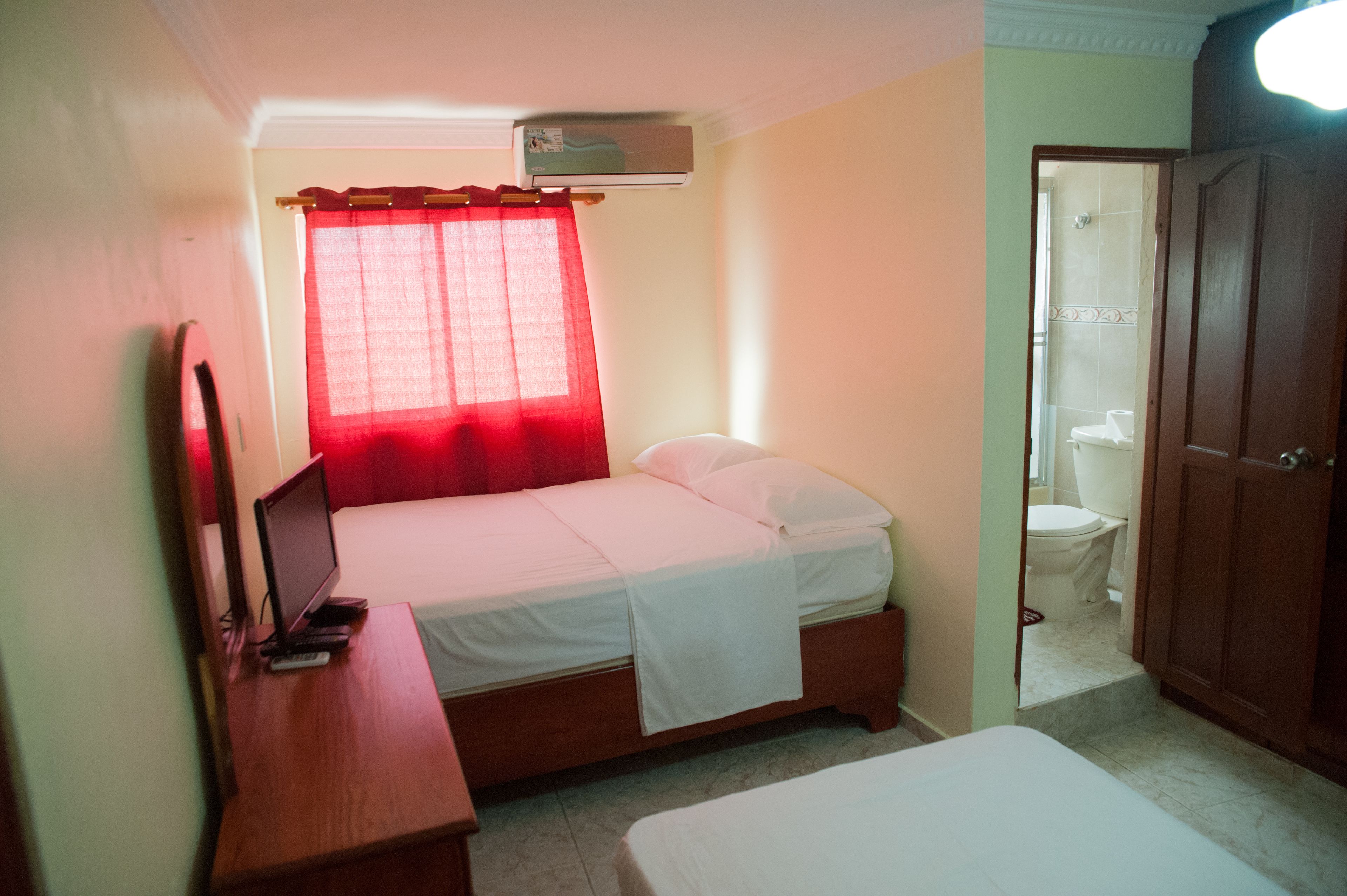 Un dormitorio con una cama, un escritorio con una computadora portátil, una ventana con cortinas rojas y una puerta que da a un baño.