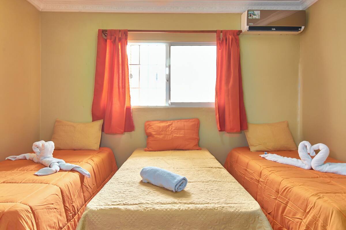 Ein Hotelzimmer mit zwei Betten, orangefarbener Bettwäsche, einem Bett mit einem zusammengerollten Handtuch und einem Fenster mit orangenen Vorhängen.