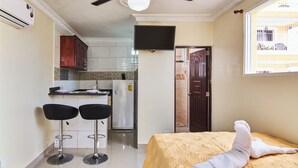 A compact hotel room with a bed, a small kitchenette, two bar stools, and a TV mounted on the wall. - Tropical Island Aparthotel (Santo Domingo Este)