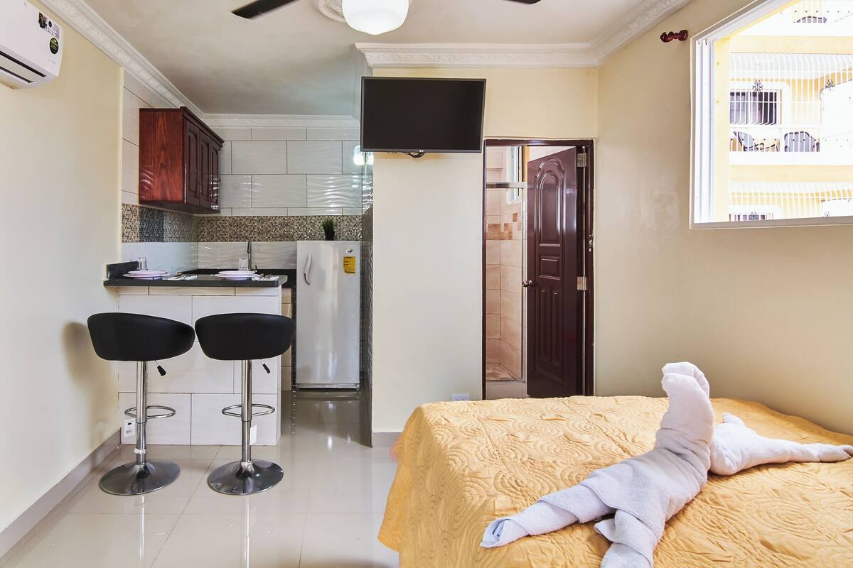 Ein kompaktes Hotelzimmer mit Bett, einer kleinen Küchenzeile, zwei Barhocker und einem an der Wand montierten Fernseher.