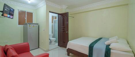 Quarto com cama, sofá vermelho, frigorífico e casa de banho visível por uma porta aberta.