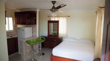 Habitación de hotel con cama, una kitchenette pequeña, un taburete de bar verde, un ventilador de techo y un espejo de baño.