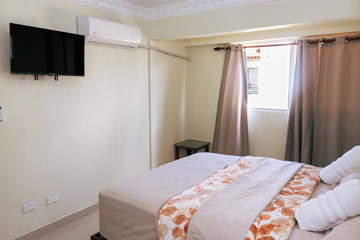 Ein Hotelzimmer mit einem Bett, einem wandmontierten Flachbildfernseher, einer Klimaanlage, einem kleinen Tisch und einem Fenster mit Vorhängen.