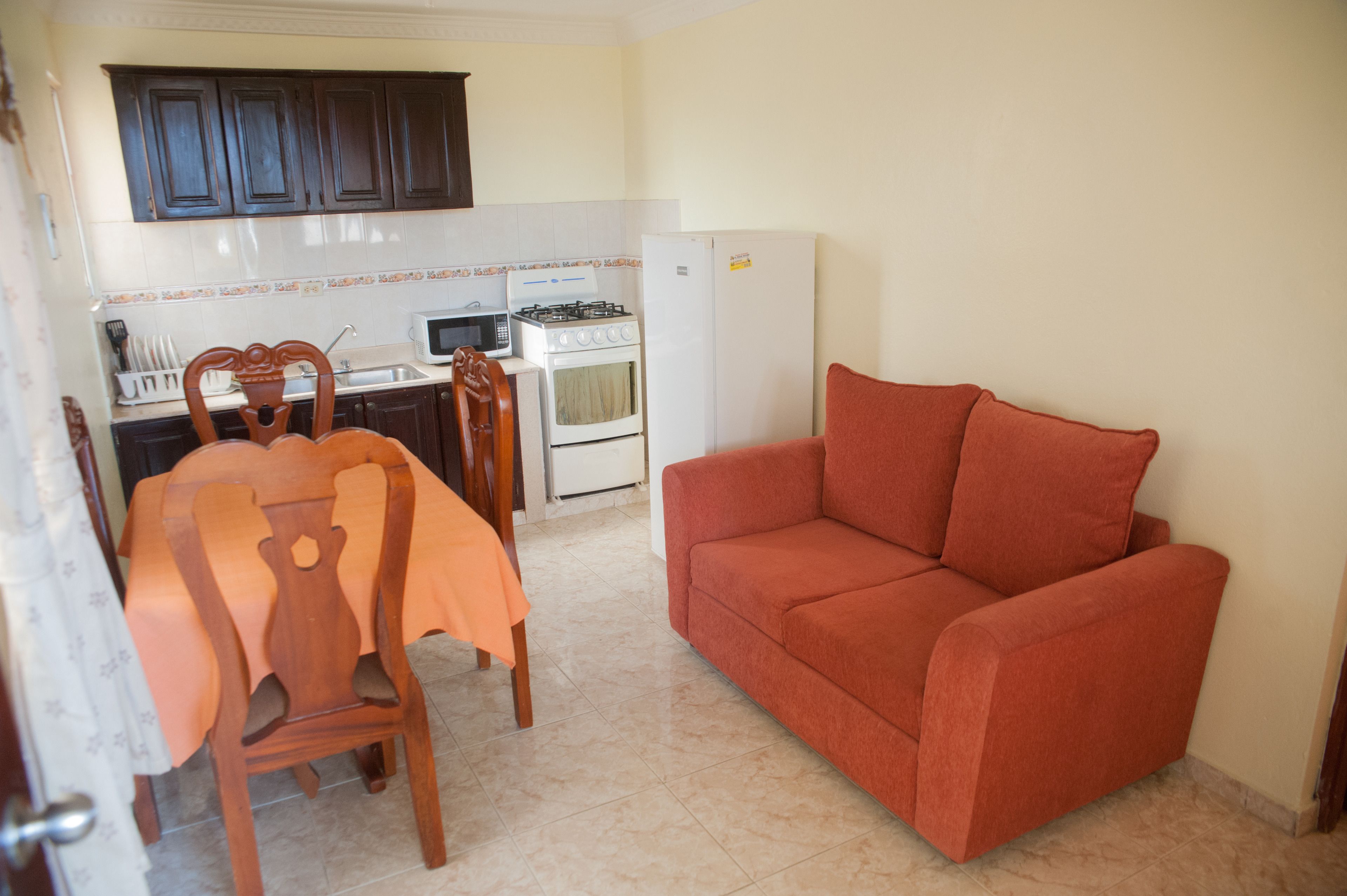 A compact kitchen with a dining table, chairs, a red sofa, and a refrigerator.