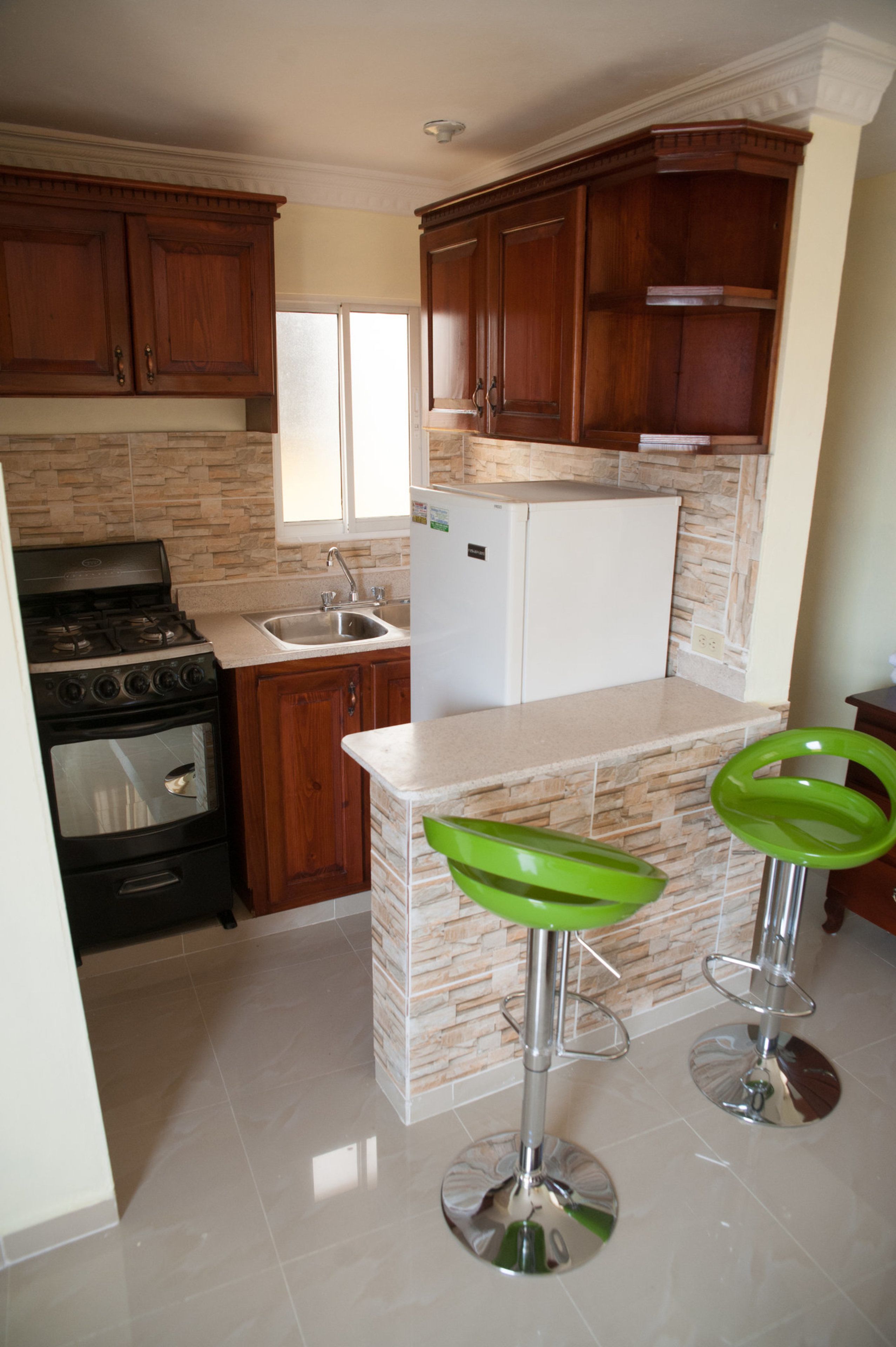 A kitchen with wooden cabinets, a white refrigerator, a stove, and a sink. There are two green bar stools.