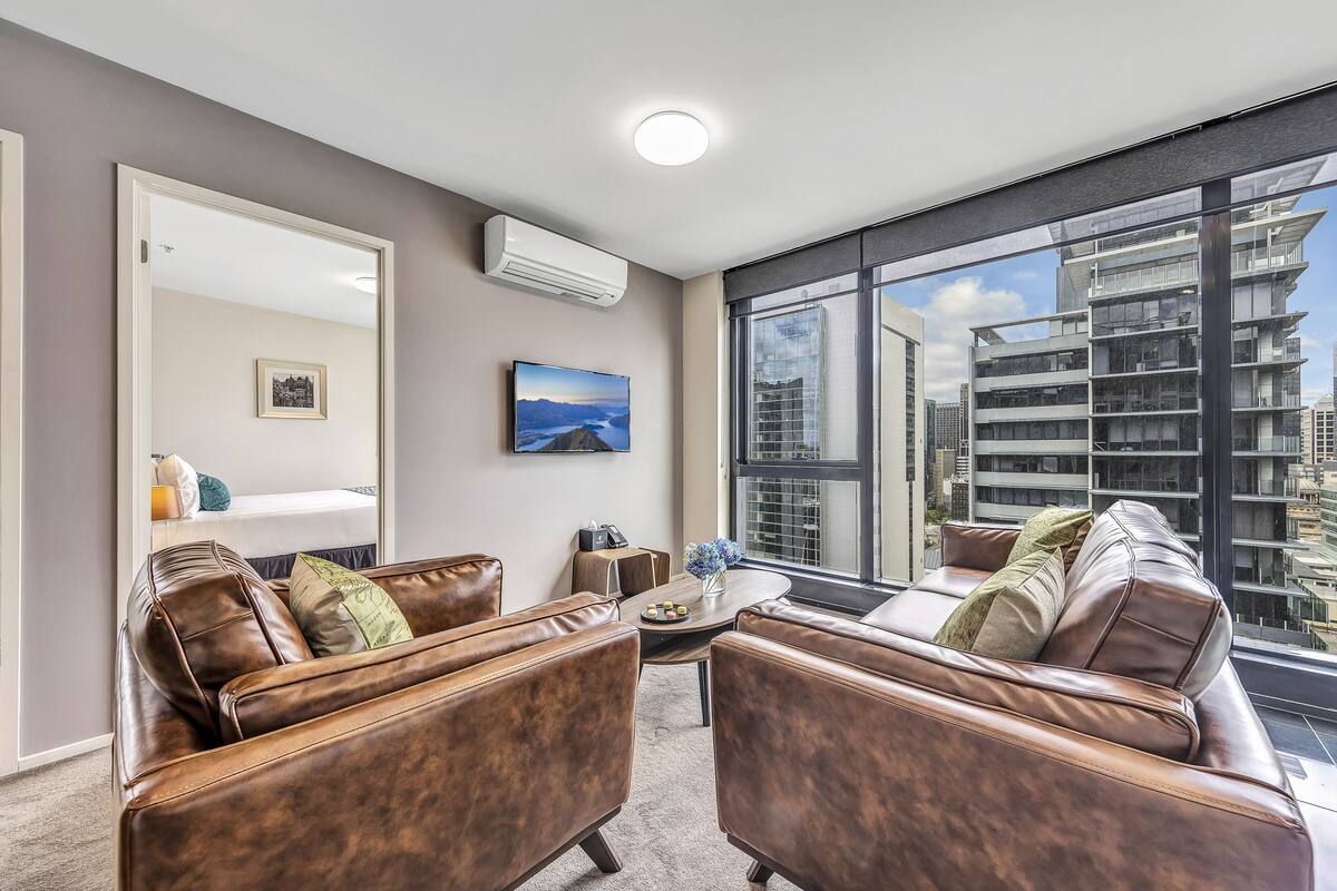 A modern living room with leather sofas, a coffee table, and a large window offering a city view.