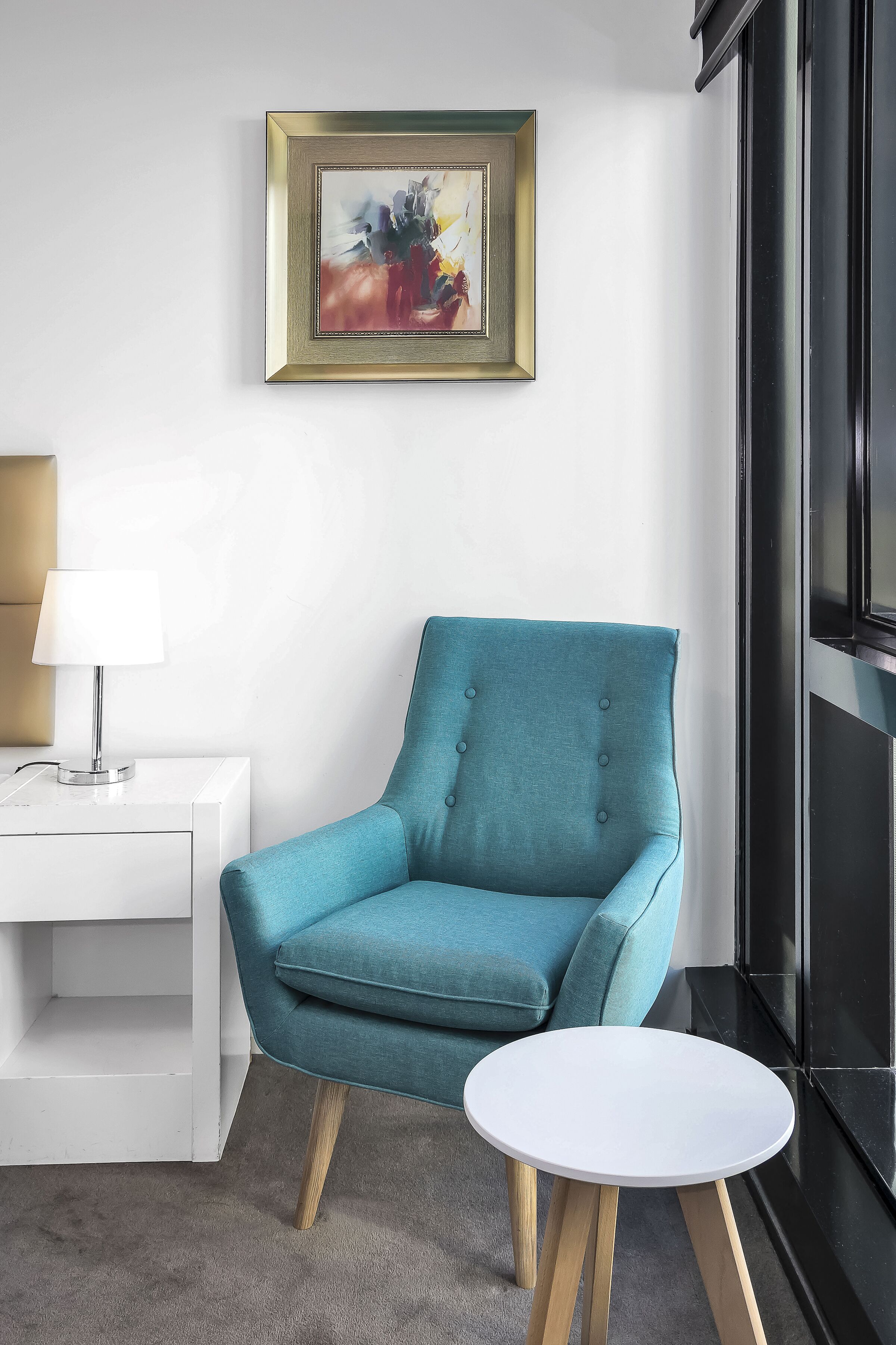 Un sillón azul verdoso con patas de madera, una mesita auxiliar blanca con una lámpara, y un cuadro abstracto enmarcado en la pared.