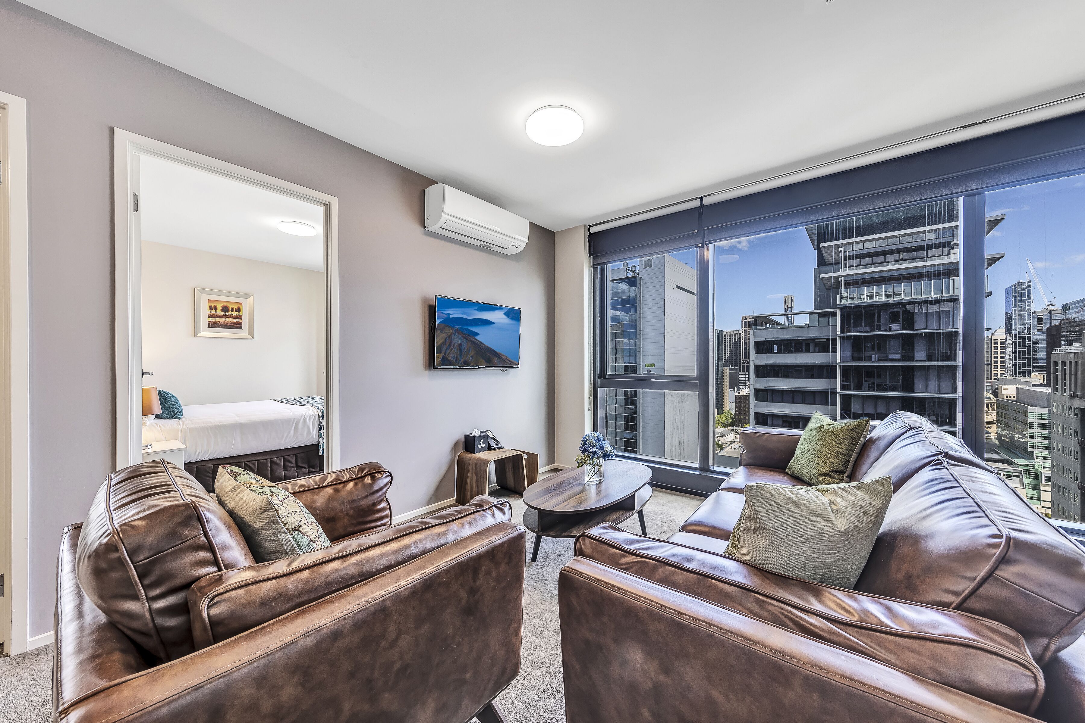 A modern living room with a large window, leather sofas, a small table, and a bed in the background.