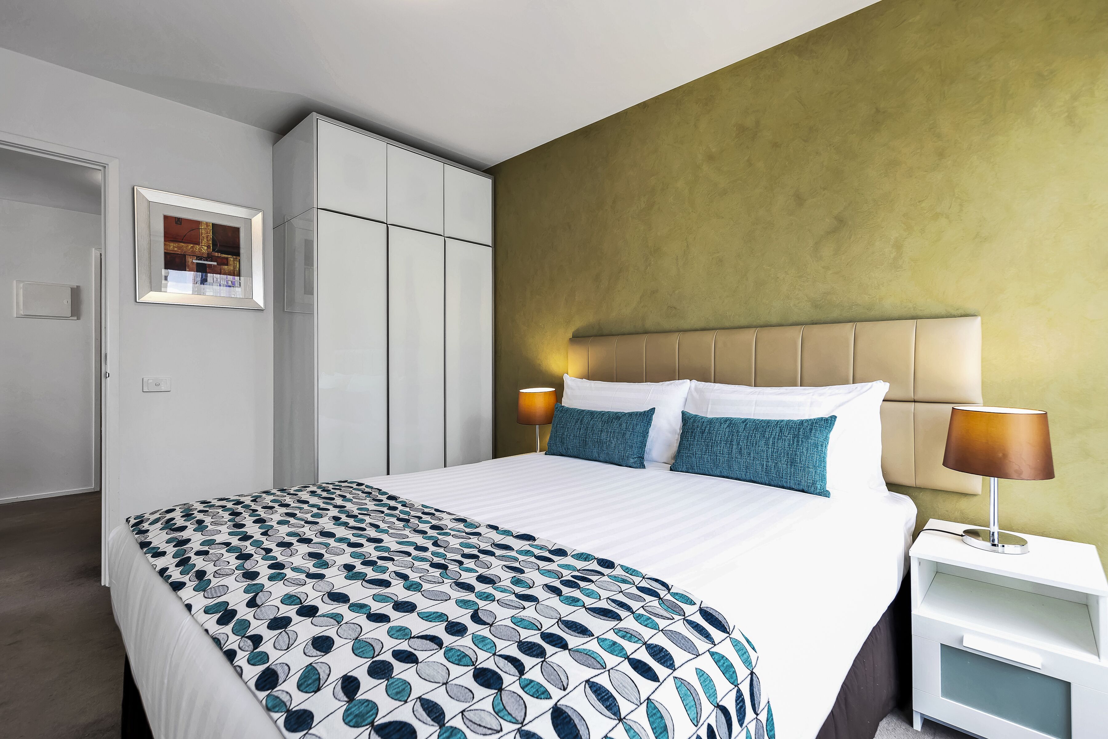 Habitación de hotel con cama, mesita de noche con lámpara, armario y un cuadro en la pared.