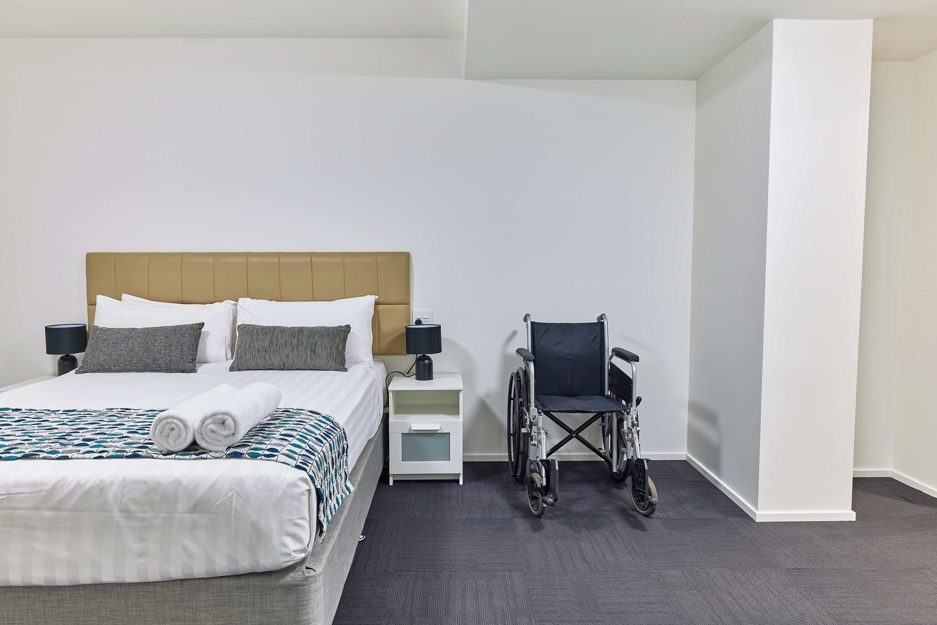 A hotel room with a bed, a wheelchair, a nightstand, and a lamp.