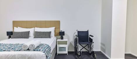 A hotel room with a bed, a wheelchair, a nightstand, and a lamp.