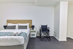 Habitación de hotel con cama, silla de ruedas, mesita de noche y lámpara.