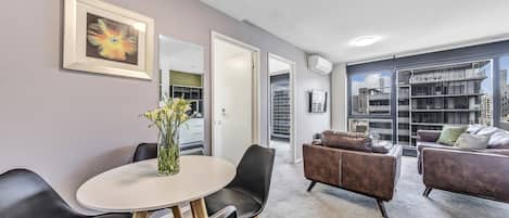 A modern living room with a dining area, a large window with a city view, and a framed artwork on the wall.