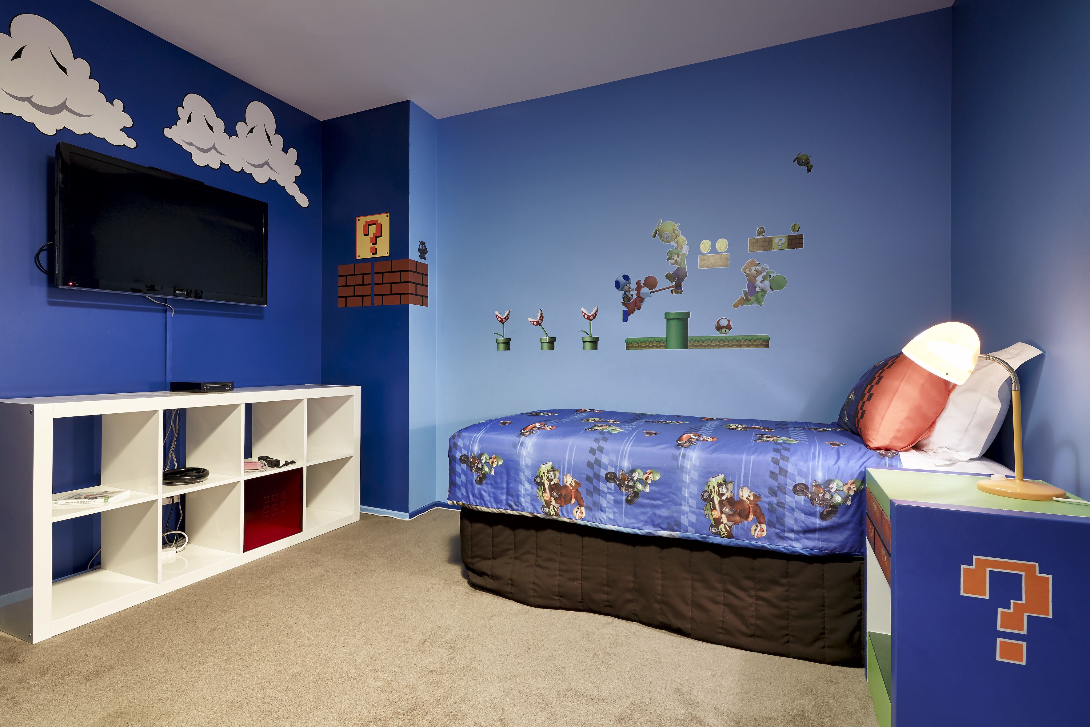 Een slaapkamer met een blauwe wand, een bed met een Mario-bedovertrek, een witte tv-meubel en een muurdecoratie van Mario-personages.